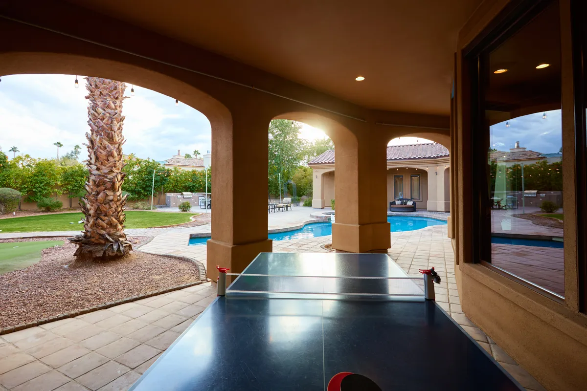 An indoor-outdoor entertainment area with a ping pong table set beside a sparkling swimming pool, framed by palm trees and a tranquil patio area. The scene exudes a playful yet serene vibe ideal for group gatherings.