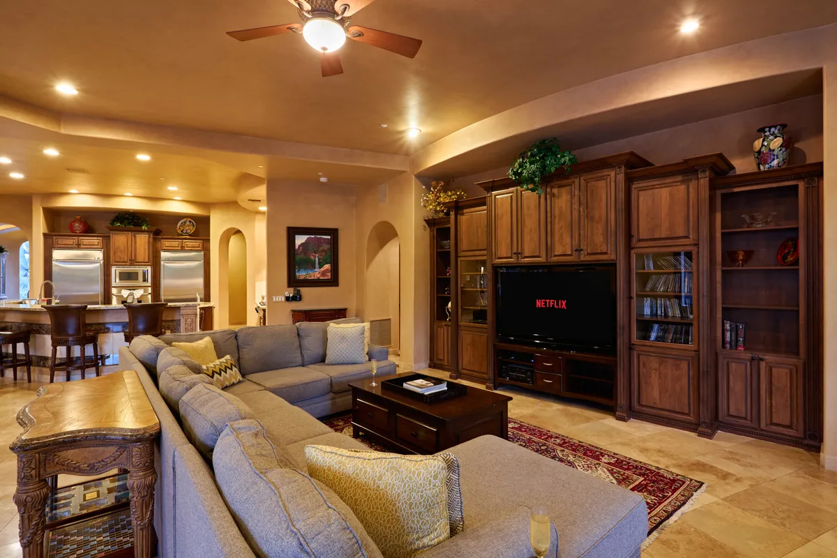 This image showcases a spacious living area with a large sectional sofa centered around an entertainment center featuring a TV, with a coffee table and a glimpse of the kitchen area in the background. The room is filled with natural light from large windows, and the open layout encourages group relaxation, movie nights, and easy flow to the kitchen.
