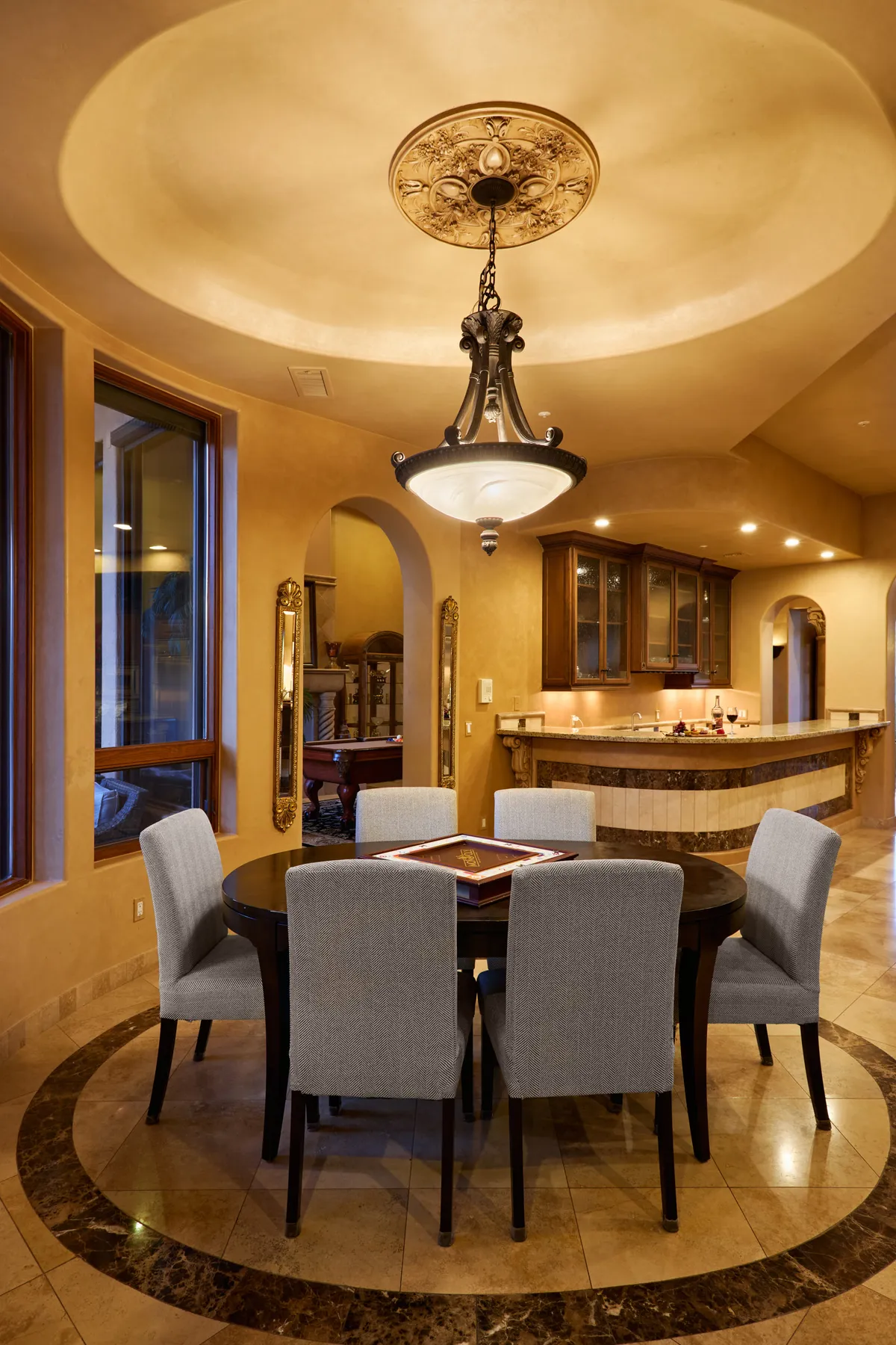 A sophisticated and warm dining area featuring a large table under an ornate chandelier, framed by expansive windows that flood the room with natural light. The setting is perfect for celebratory feasts and intimate gatherings, with elegant décor and plush seating.