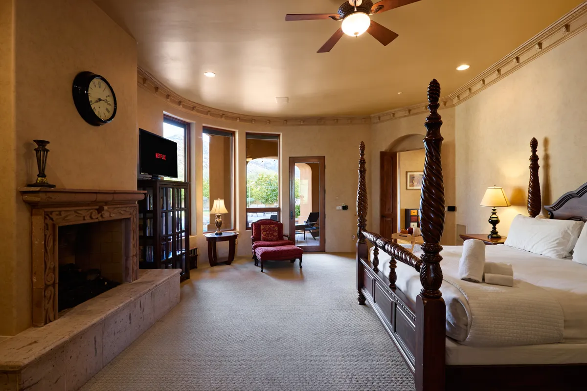 This image showcases a spacious bedroom featuring a four-poster bed, a cozy fireplace, large windows letting in natural light, a comfortable armchair, and a television. The arrangement radiates comfort and luxury, aligning with the concept of a restful sanctuary.