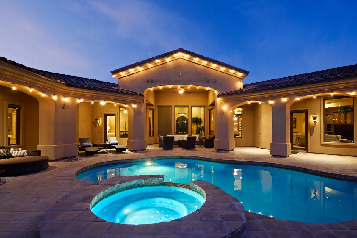 A sparkling swimming pool is surrounded by comfortable outdoor seating, all softly illuminated by string lights. The inviting pool deck is set for evening gatherings, with plenty of space for guests to lounge and socialize. The adjacent hot tub and outdoor kitchen area, while visually present, are subtly set in the background.