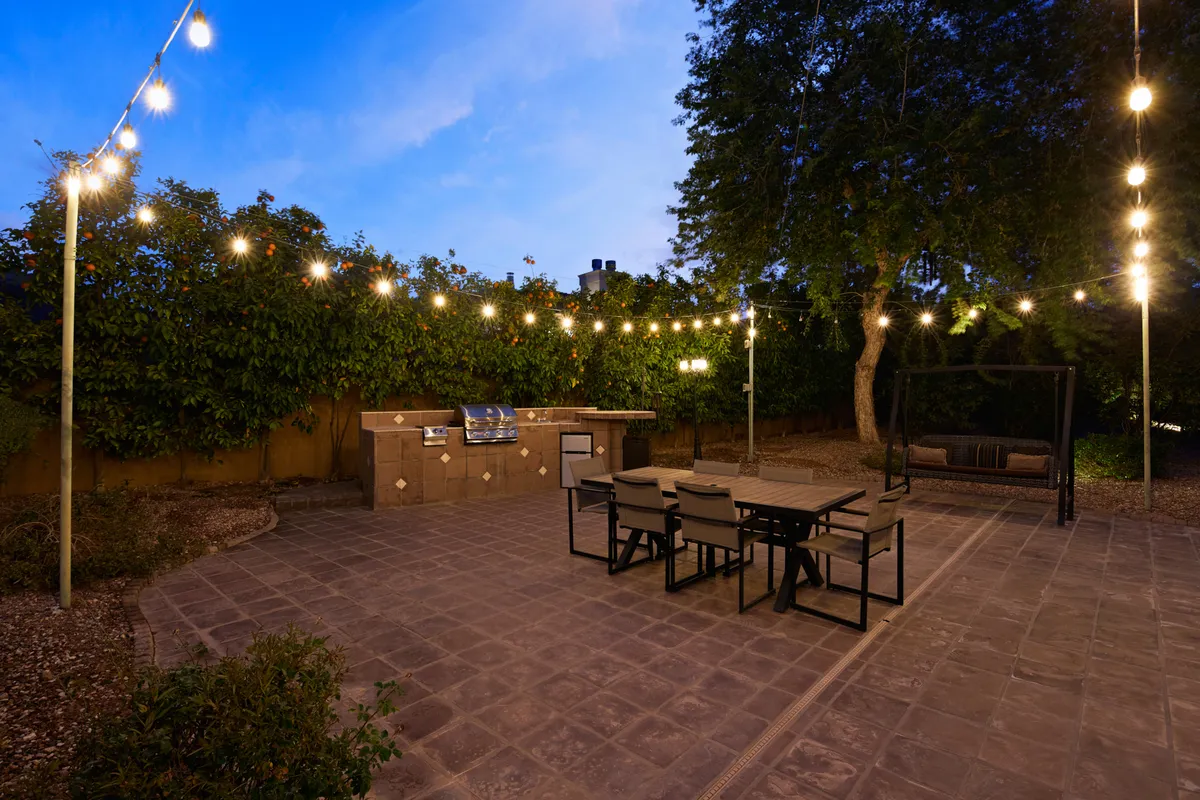 The outdoor kitchen and dining area are highlighted here, with a modern BBQ setup under string lights, a spacious dining table for al fresco meals, and a charming swing chair. The scene is warmly lit, suggesting a welcoming space for barbecues and evening gatherings.
