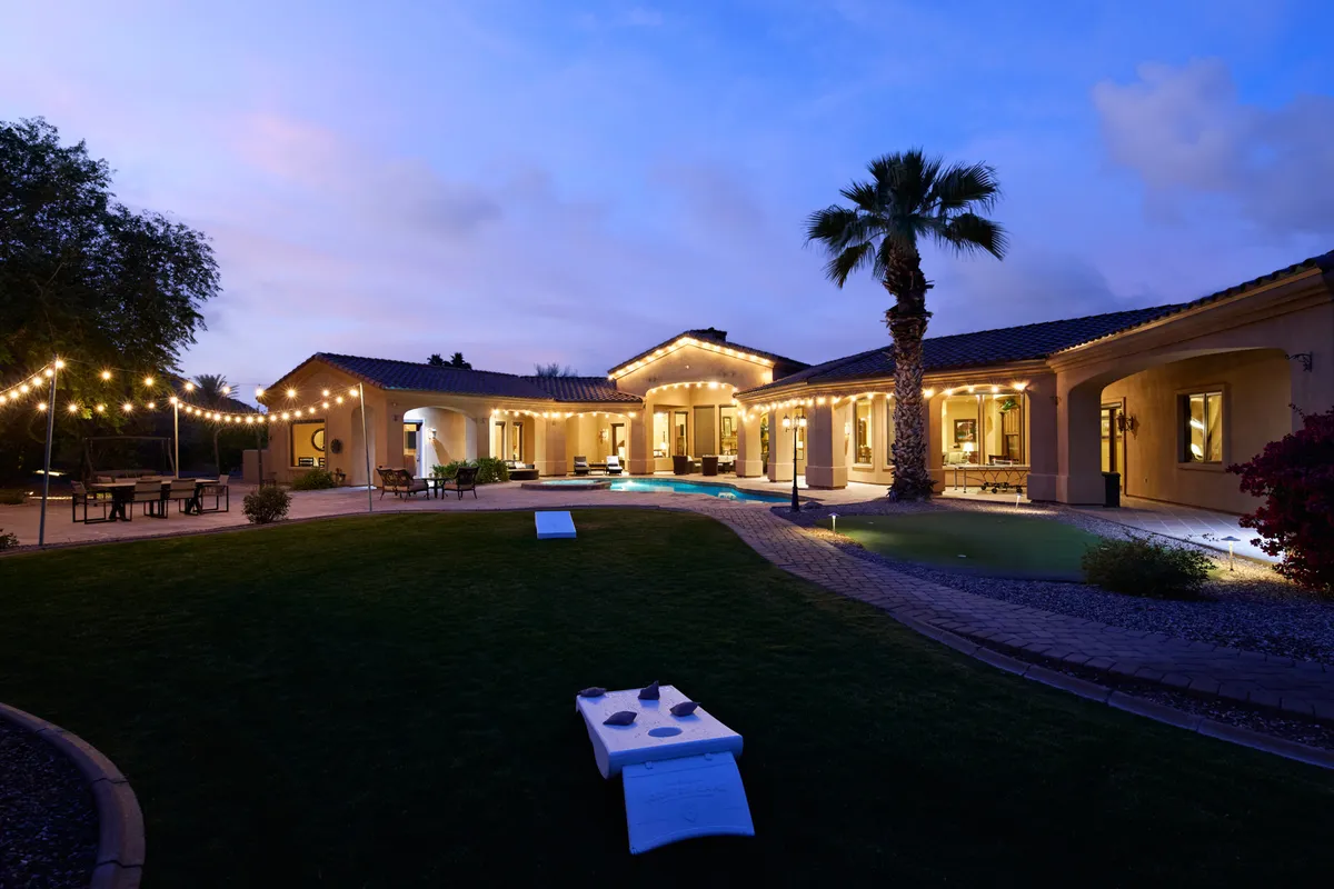 The estate’s illuminated facade glows warmly at dusk, with the pool, palm trees, and outdoor lounge areas visible in the foreground. String lights enhance the festive mood, creating an inviting setting for al fresco dining, lively games, and mingling with friends.