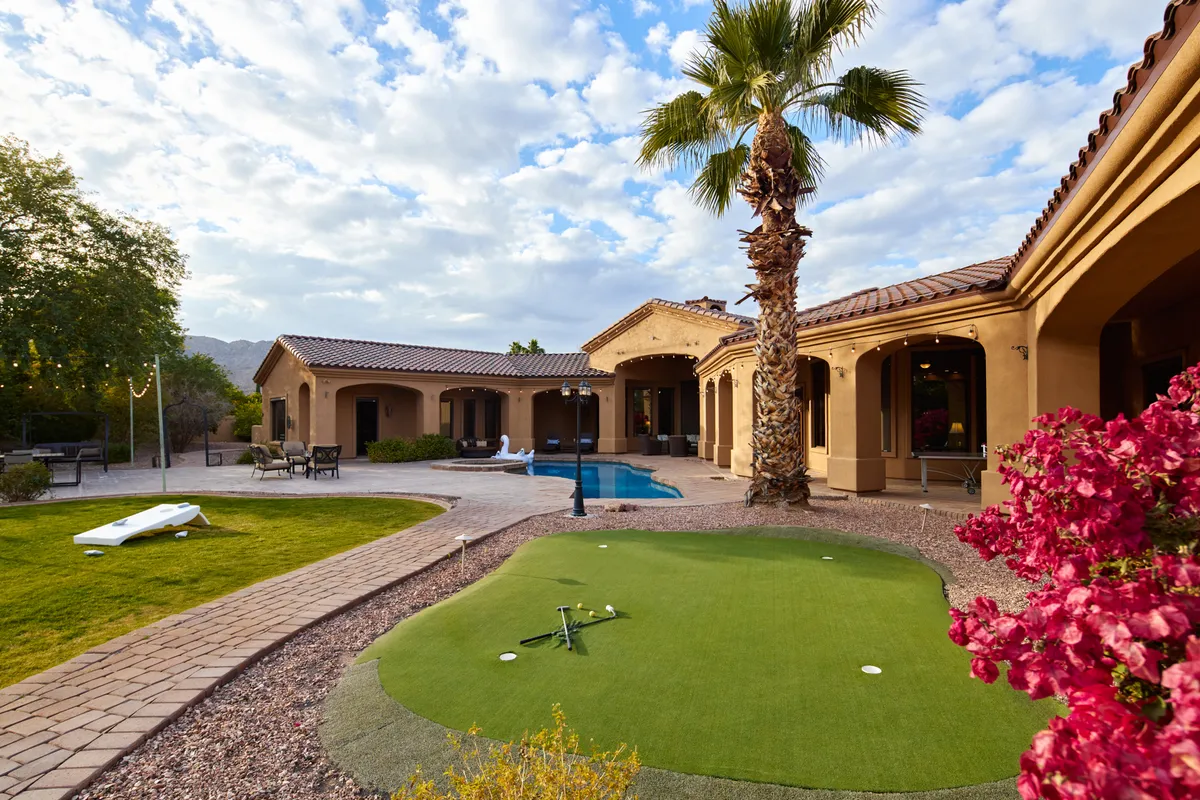 A view of the backyard featuring a sparkling swimming pool, lush landscaping, a private putting green, and a palm tree. The scene is set for outdoor fun and relaxation, with spaces for swimming, socializing, and playing games together.