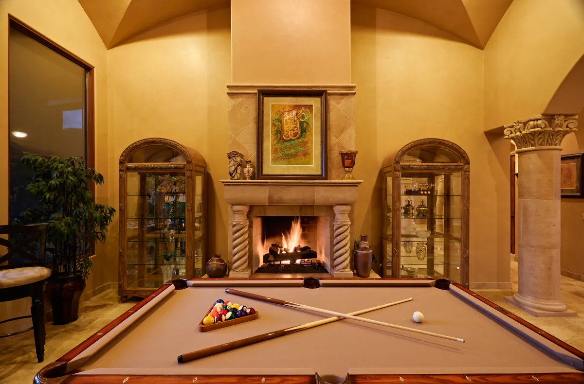 A stylish, relaxed game area with a pool table at the center, flanked by a cozy fireplace and tasteful artwork adorning the wall. Display cabinets and seating suggest a space designed for both friendly competition and socializing.